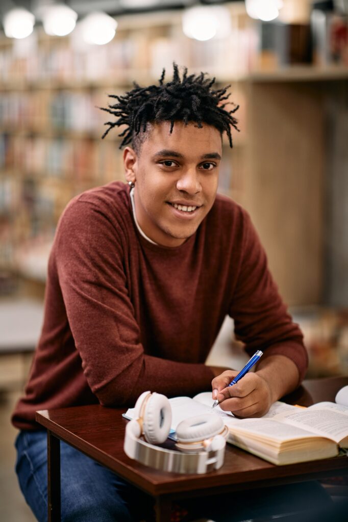 Young black man taking notes during a research in a library and looking at camera.